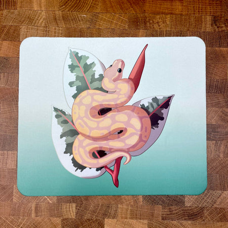 A mousepad decorated with an illustration of a yellow and peach colored ball python snake, known as a banana ball, in front of a plant on a green background.