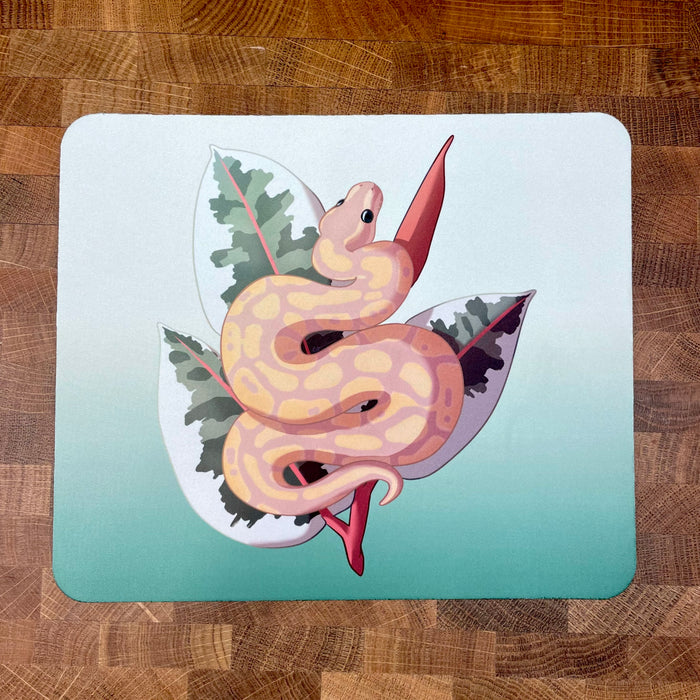 A mousepad decorated with an illustration of a yellow and peach colored ball python snake, known as a banana ball, in front of a plant on a green background.