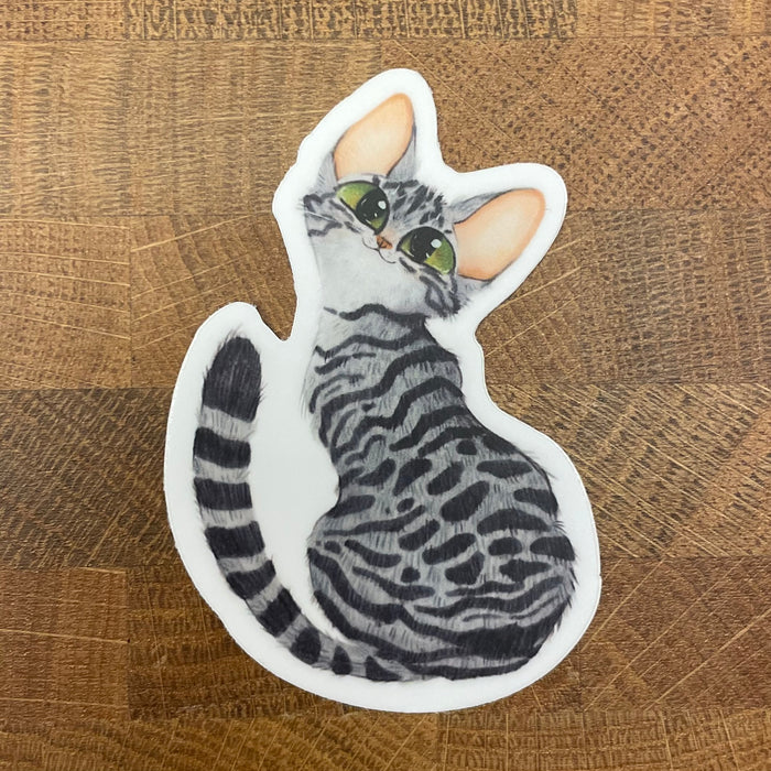 Die cut sticker of a grey tabby cat with green eyes photographed on a wooden surface