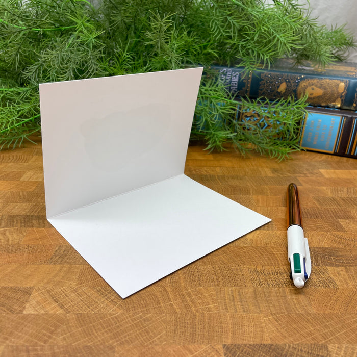 White, blank greeting card on a wooden surface with greenery and books in the background