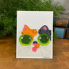 Card with a colorful cat face design on a wooden surface with greenery in the background