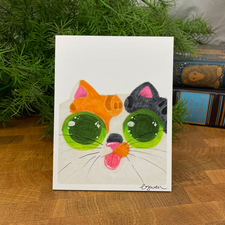 Card with a colorful cat face design on a wooden surface with greenery in the background
