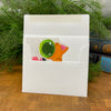 White card envelope with a colorful card featuring a cat design, placed on a wooden surface with green plants in the background.