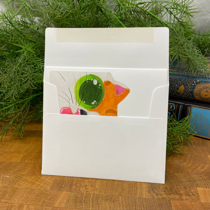 White card envelope with a colorful card featuring a cat design, placed on a wooden surface with green plants in the background.