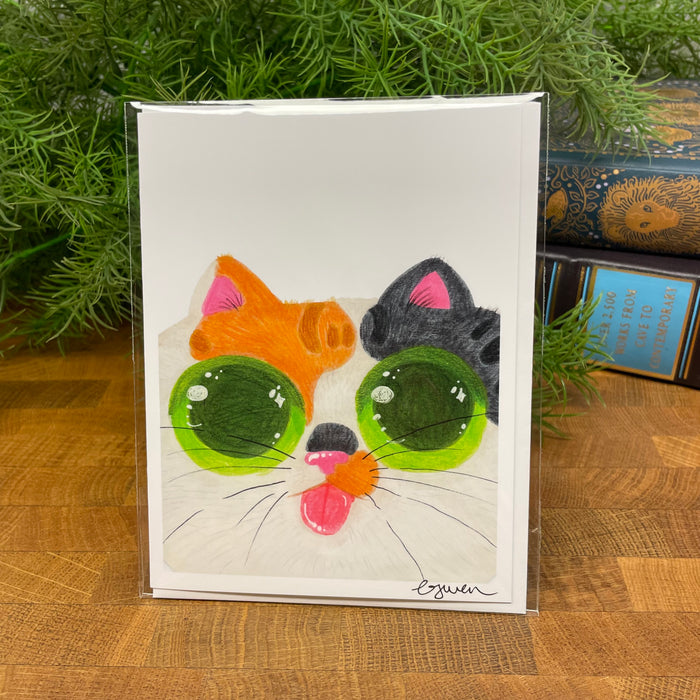 Card with a colorful cat face design and a white envelope packaged in a clear bag, on a wooden surface with greenery in the background