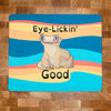 A mousepad with a realistic cartoon style illustration of a crested gecko, sitting down, looking forward while licking one eye with the caption, "Eye Lickin' Good" on a colorful background, the pun and the art are created by Studio Zoologic.