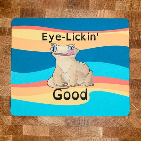 A mousepad with a realistic cartoon style illustration of a crested gecko, sitting down, looking forward while licking one eye with the caption, "Eye Lickin' Good" on a colorful background, the pun and the art are created by Studio Zoologic.