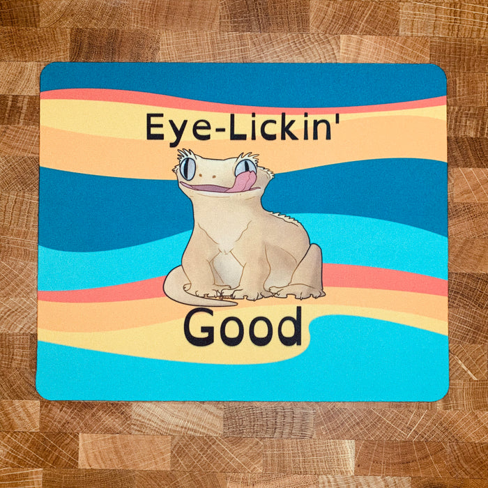 A mousepad with a realistic cartoon style illustration of a crested gecko, sitting down, looking forward while licking one eye with the caption, "Eye Lickin' Good" on a colorful background, the pun and the art are created by Studio Zoologic.