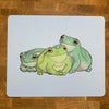 A mousepad decorated with an illustration of three green tree frogs on a light blue background.